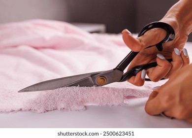 A close-up of a person's hand using large scissors to cut a piece of pink fabric on a white surface. The focus is on the scissors and the fabric, showcasing a crafting or sewing activity.