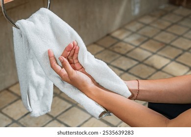 A close-up of a person's hands drying with a white towel in a bathroom setting. The background features a wooden towel rack and tiled walls, creating a clean and modern atmosphere.