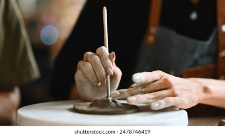 A close-up of a person's hands shaping clay on a pottery wheel, with one hand holding a tool and the other hand sculpting. The background is blurred, highlighting the focus on the hands and the clay. - Powered by Shutterstock