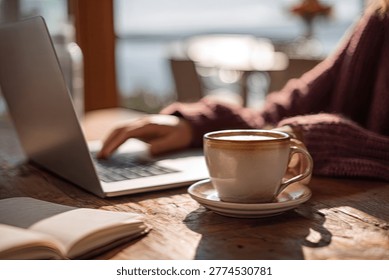 Close-up of a person's hands typing on a laptop, with a cup of coffee and an open book on a wooden table.
