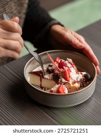 Close-up of a person's hands using a silver fork to eat a slice of creamy Basque burnt cheesecake, beautifully garnished with fresh strawberries, blueberries, and red berry sauce in a gourmet restaura