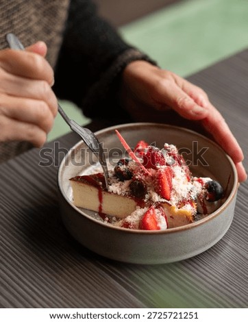 Close-up of a person's hands using a silver fork to eat a slice of creamy Basque burnt cheesecake, beautifully garnished with fresh strawberries, blueberries, and red berry sauce in a gourmet restaura