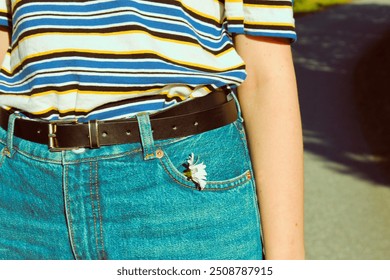 Close-up of a person's waist wearing a striped shirt, blue jeans with a black belt, and a small white flower tucked in the front pocket. - Powered by Shutterstock