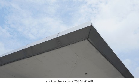 Close-up perspective of a contemporary building roof angular design contrasting against a light blue sky with scattered clouds, emphasizing modern architectural aesthetics and urban structural element
