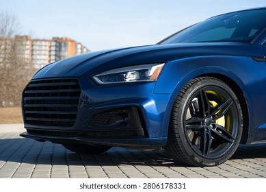 A close-up perspective of the front driver's side. The shot highlights the black multi-spoke alloy wheel paired with a striking yellow brake caliper. Also visible are the LED headlight, the edge of the black front grille, and the deep blue metallic reflections on the fender.