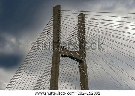 Close-up perspective of a massive concrete cable-stayed bridge pylon supporting numerous thick steel suspension cables against a moody, overcast, and dramatic sky