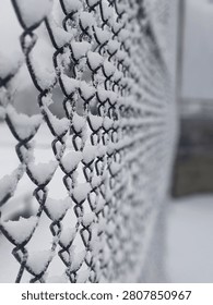 A close-up perspective shot looking down a metal chain-link fence covered in fresh white snow. The shallow depth of field heavily blurs the background on a cold winter day.