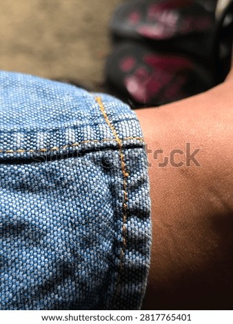 A close-up perspective showcases the textured surface of a light blue woven denim fabric with a prominent stitched seam running along the center. The selective focus accentuates the fine details of the yellow contrast thread, emphasizing the strength and precision of industrial garment assembly. The woven cotton fibers display a distinct pattern of alternating light blue and off-white yarns, giving the material an authentic, tactile appearance. The shallow depth of field causes the upper and lower portions of the image to softly blur into a warm, neutral background, making the sharp central stitching stand out dramatically. This detailed photograph highlights the artistry of clothing construction, tailor craftsmanship, and durable textile manufacturing. It offers an excellent visual asset for fashion design, textile texture references, casual wear promotions, apparel production guides, and material studies. The dynamic angle adds a sense of depth and dimension to the classic indigo fabric structure.
