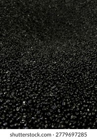 Close-up perspective of thousands of tiny, uniform black plastic pellets (nurdles) piled high in an industrial storage setting, showcasing the raw material for injection molding and plastic manufacturing
