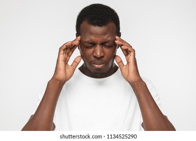 Closeup Photo Of African American Man Isolated On Grey Background Dressed In White T-shirt Pressing Fingers Of Both Hands To Temples Experiencing Great Headache That Prevents Him From Thinking