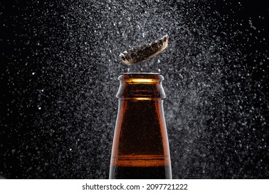 Closeup photo of an amber beer bottle splashing beer drops on a black background. Beer cap flying on top of the bottle.