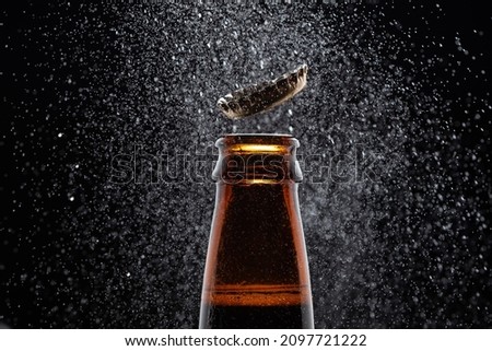 Closeup photo of an amber beer bottle splashing beer drops on a black background. Beer cap flying on top of the bottle.