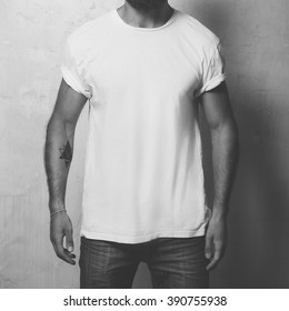 Closeup Photo Bearded Angry Man With Tattoo Wearing White Blank T-shirt And Black Jeans, Standing Opposite Concrete Empty Wall. Vertical ,mockup