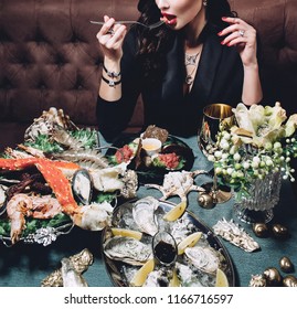 close-up photo of a beautiful woman sitting in a black jacket and expensive jewelry, red manicure at a table teeming with fresh seafood in a restaurant, and puts a piece of tuna in her mouth with fork