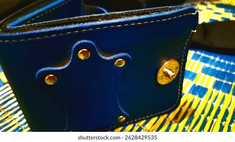 A close-up photo of a blue leather wallet with golden metal studs and a snap button. The wallet is placed on a woven mat with a blue and yellow pattern. The stitching and texture of the leather