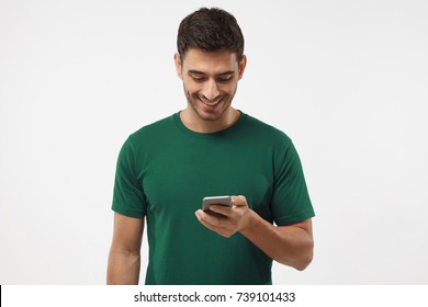 Closeup photo of casually dressed student standing isolated on gray background looking at screen of smart phone, browsing web pages and smiling while chatting with friends - Powered by Shutterstock