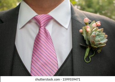Close-up photo of a Caucasian male wearing a dark gray suit, collared white shirt and a pink neck tie with diagonal stripes. Pinned to his suit is a boutonniere with tiny pink flowers and a succulent.