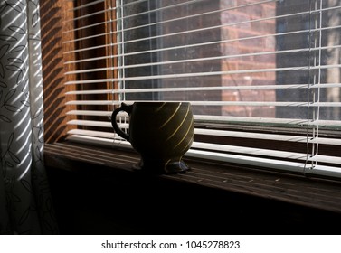 A Closeup Photo Of Coffee Mug Sitting On Window Ledge. The Window Blinds Are Open, Creating A Line Of Shadows On The Mug.