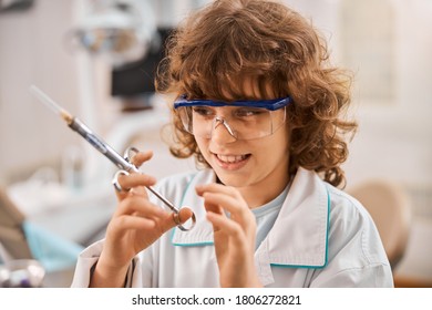 Close-up Photo Of A Curly Kid With A Sneaky Smile Wearing A Lab Coat Holding A Medical Syringe And Looking Cunning