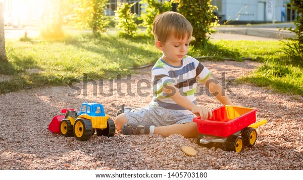 Closeup photo of cute little boy\
playing on the palyground with toys. Child having fun with truck,\
excavator and trailer. He is pretending to be a builder or\
driver
