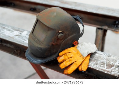 Closeup photo of a dusty black welding helmet and yellow protective work gloves on metal surface in a factory. - Powered by Shutterstock