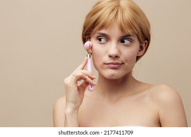 Close-up Photo. An Emotional Woman In A Towel With Clean, Beautiful, Well-groomed Skin, Short Red Hair On A Beige Background Does A Facial Massage With A New Massage Roller. Horizontal Studio Shot.