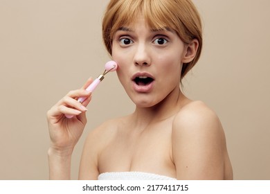 Close-up Photo. An Emotional Woman In A Towel With Clean, Beautiful, Well-groomed Skin, Short Red Hair On A Beige Background Does A Facial Massage With A New Massage Roller. Horizontal Studio Shot.