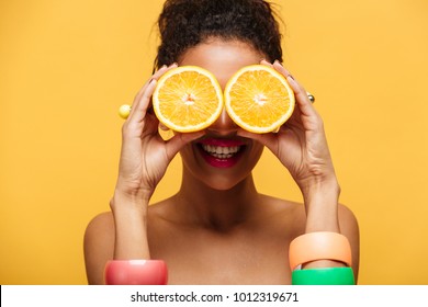 Closeup photo of funny mixed-race woman with fashion accessories having fun and covering eyes with two halves of orange isolated over yellow wall