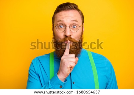 Closeup photo of funny red head guy tricky mood hold finger on lips asking not talk say speak tell wear specs bright blue shirt green suspenders isolated yellow color background