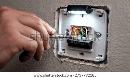 Close-up photo of a hand using a precision screwdriver to secure wires onto a thermostat terminal block during installation or repair, illuminated by an integrated light.