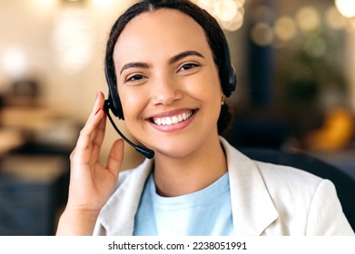 Close-up photo of a happy successful brazilian or latino young woman, with headset, call center agent, support service operator, sits in the office, consult clients online, looks at camera, smiling