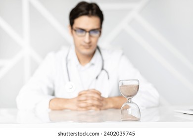 Close-up photo of hourglass on the desktop and male doctor on background. Time and health concept