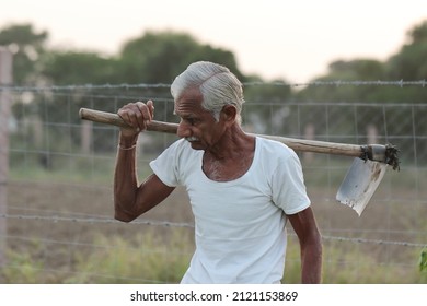 Close-up Photo Of An Indian Senior Male Farmer Man Works In The Field With A Shovel On His Shoulder With Wearing A White Vest