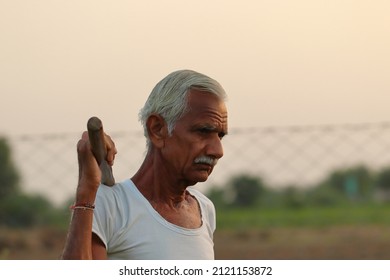 Close-up Photo Of An Indian Senior Male Farmer Man Stands In The Field At Sunset With A Shovel On His Shoulder With Wearing A White Vest