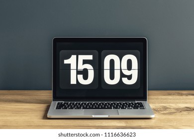 Close-up Photo Of Laptop With Time Screensaver Placed On Wooden Desk In Room Interior With Dark Wall