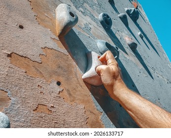 Close-up Photo Of Male Hand Gripping Climbing Holds On Worn Wall Outside. Artificial Rock Climbing Wall At Park.