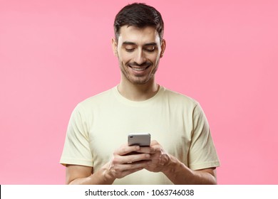 Closeup Photo Of Man In Beige Tshirt Standing Isolated On Pink Background Looking Attentively At Screen Of Cellphone, Browsing Web Pages And Smiling Nicely While Chatting With Friends