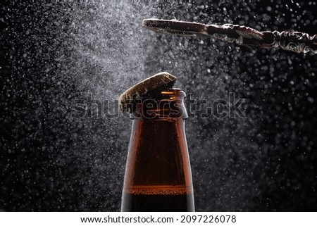 Closeup photo of a metal bottle opener opening an amber beer bottle splashing beer drops on a black background