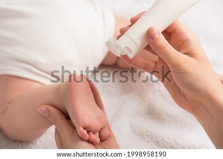 Closeup photo of newborn laying on stomach and mother's hands holding baby's foot and white cream tube on isolated white textile background