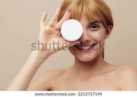 Close-up photo. A nice woman in a towel with clean skin, a beautiful smile and red hair pinned back hair on a beige background with a jar of cream in her hand.Horizontal studio shot.