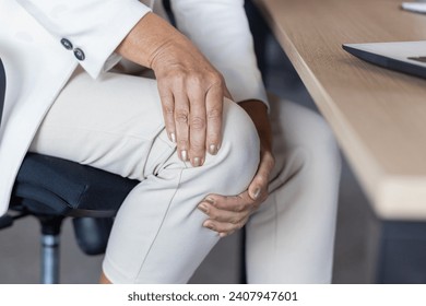 Close-up photo in the office at the desk. The hands of an older woman in a business suit are holding the knee of the leg, giving a massage. - Powered by Shutterstock