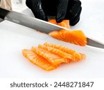 A close-up photo of a person carefully slicing a fresh salmon fillet on a cutting board. The salmon is destined to be served as sashimi, a Japanese dish of thinly sliced raw fish.