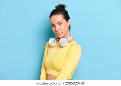 Closeup photo portrait of sporty girl model wearing yellow yoga top and white headphones on her neck, girl model posing on studio blue backdrop, natural beauty concept, copy space, high quality photo
