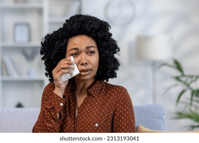 Close-up photo. Portrait of upset young African American woman sitting on sofa at home and crying, suffering from depression, wiping tears with napkin.