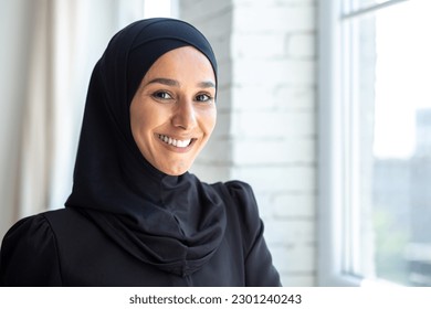 Close-up photo. Portrait of a young beautiful Muslim woman in a black hijab looking confidently and smiling at the camera, standing indoors by the window. - Powered by Shutterstock