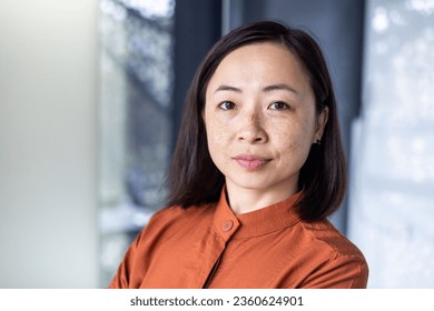 Close-up photo portrait of a young beautiful confident Asian woman standing in an office center and seriously looking at the camera. - Powered by Shutterstock