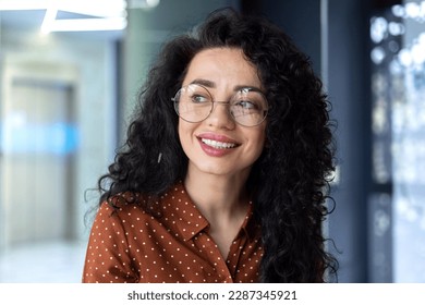 Close-up photo. Portrait of young hispanic woman with glasses and curly hair sitting, working and studying in office, campus. Smiling, she looks to the side. - Powered by Shutterstock