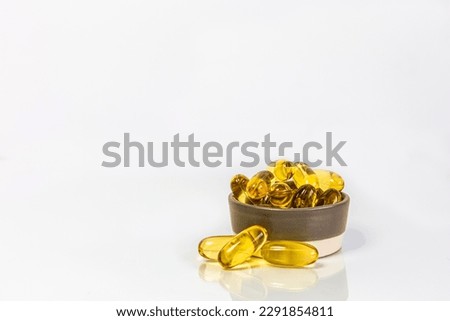 A close-up photo of several yellow capsules of fish oil in a small bowl on a white background. Copy space, reflection.