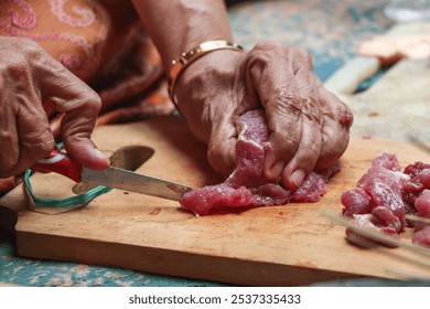 A close-up photo showing the hands of an elderly woman cutting raw red meat using a small knife on a wooden chopping board - Powered by Shutterstock