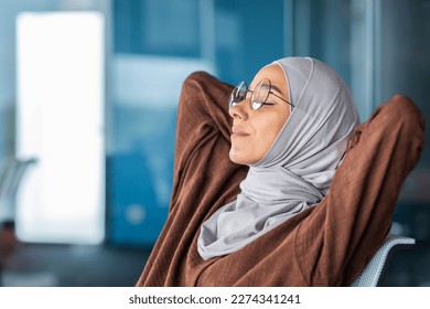 Close-up photo from the side. A tired young Arab woman in a hijab and glasses sits in the office on a chair with her hands behind her head, rests, sleeps, closes her eyes. - Powered by Shutterstock